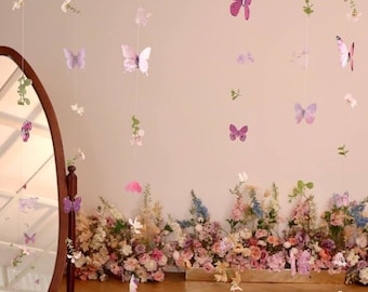 May include: A set of butterfly and flower garlands, perfect for a party or special event. The garlands are made of paper and feature a variety of colors, including pink, purple, and white. The garlands are hanging from the ceiling and are surrounded by a wall of flowers.