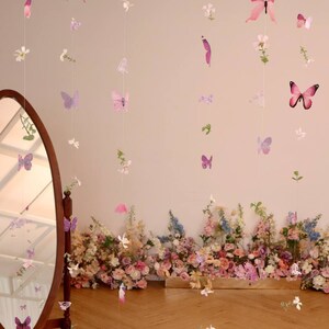 Pu&ograve; includere: Un set di ghirlande di farfalle e fiori rosa e viola appese al soffitto. Le ghirlande sono fatte di carta e sono infilate su un filo trasparente. Le ghirlande sono appese davanti a un muro bianco. Ci sono fiori in primo piano.