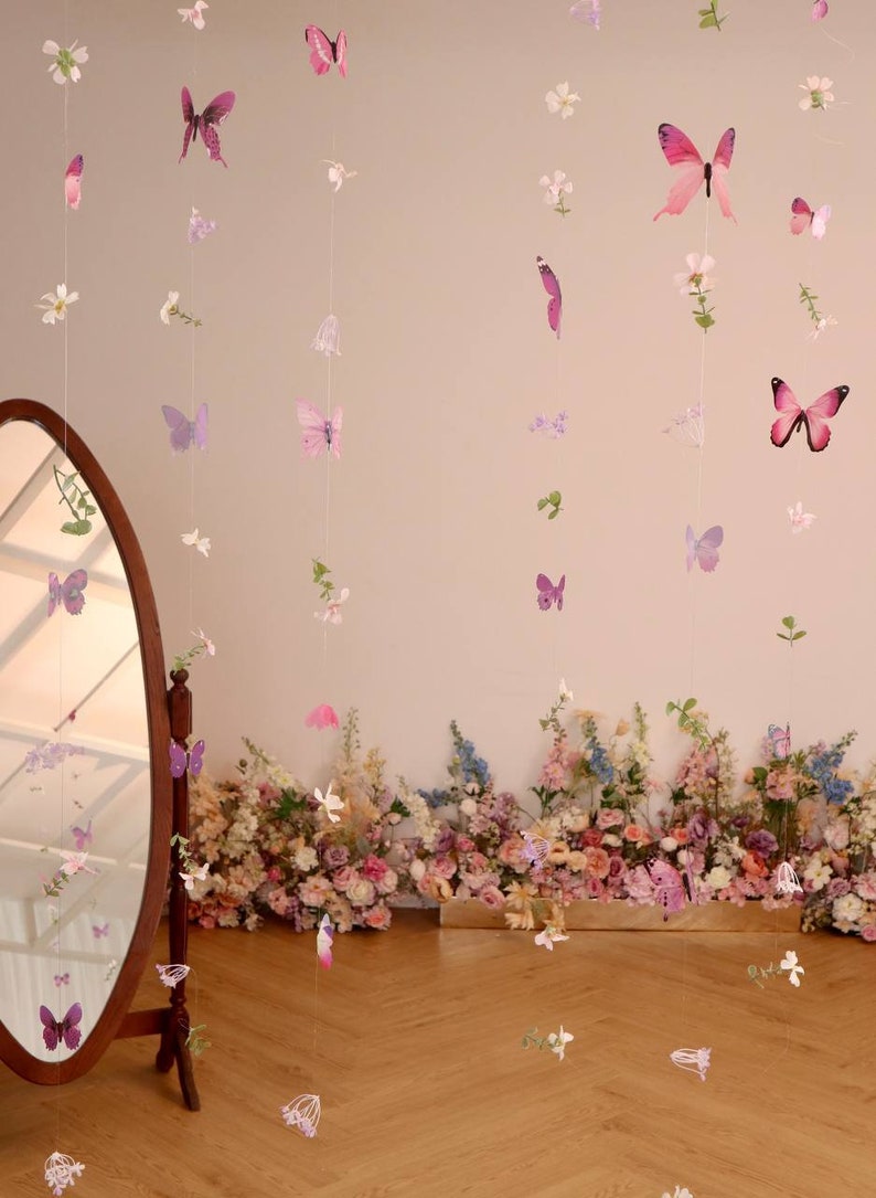 Pu&ograve; includere: Un set di ghirlande di farfalle e fiori rosa e viola appese al soffitto. Le ghirlande sono fatte di carta e sono infilate su un filo trasparente. Le ghirlande sono appese davanti a un muro bianco. Ci sono fiori in primo piano.
