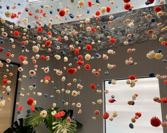 May include: A room decorated with strings of colorful paper flowers hanging from the ceiling. The flowers are in shades of pink, red, blue, and white. There is a large plant in the corner of the room and a gray couch in the foreground.