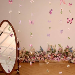 Pu&ograve; includere: Un set di ghirlande di farfalle rosa e viola con fiori bianchi appesi al soffitto. Le ghirlande sono fatte di carta e sono infilate su un filo trasparente. Le farfalle sono di diverse dimensioni e forme. Le ghirlande sono appese davanti a un muro bianco con uno specchio e un'arrangiamento floreale.