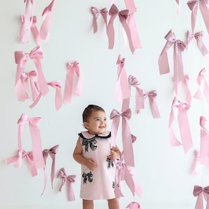 Könnte beinhalten: Ein lächelndes Kleinkind in einem rosa Kleid mit schwarzen Schleifenakzenten und einem schwarzen Kragen steht inmitten hängender rosa Schleifen. Die Schleifen in verschiedenen Größen und Rosatönen schaffen eine verspielte Atmosphäre.