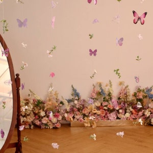Pu&ograve; includere: Un set di ghirlande di farfalle e fiori rosa e viola appese al soffitto. Le ghirlande sono fatte di carta e sono infilate su un filo trasparente. Le ghirlande sono appese davanti a un muro decorato con un'arrangiamento floreale.