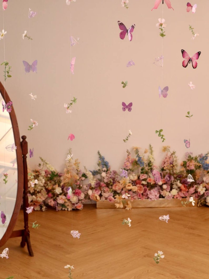 Pu&ograve; includere: Un set di ghirlande di farfalle e fiori rosa e viola appese al soffitto. Le ghirlande sono fatte di carta e sono infilate su un filo trasparente. Le ghirlande sono appese davanti a un muro decorato con un'arrangiamento floreale.