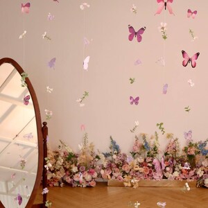 Pu&ograve; includere: Un muro bianco con una ghirlanda di farfalle e fiori rosa e viola appesa. La ghirlanda &egrave; fatta di carta ed &egrave; infilata su uno spago trasparente. C'&egrave; uno specchio sul lato sinistro dell'immagine e un'arrangiamento floreale in basso.