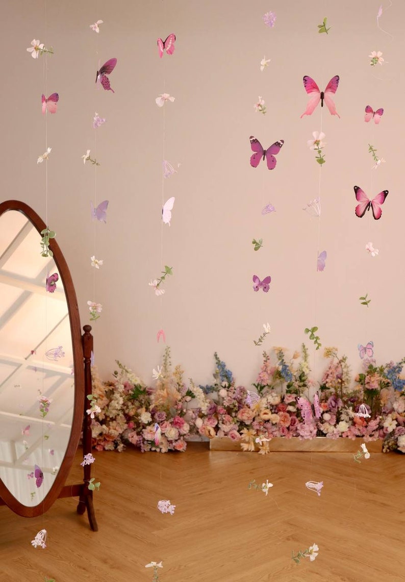 Pu&ograve; includere: Un muro bianco con una ghirlanda di farfalle e fiori rosa e viola appesa. La ghirlanda &egrave; fatta di carta ed &egrave; infilata su uno spago trasparente. C'&egrave; uno specchio sul lato sinistro dell'immagine e un'arrangiamento floreale in basso.