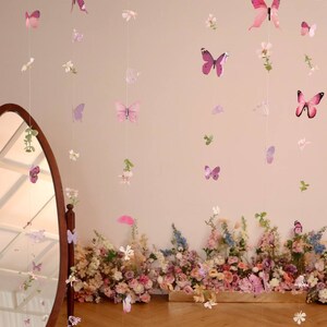 Pu&ograve; includere: Un muro rosa con una ghirlanda floreale con farfalle appese. La ghirlanda &egrave; fatta di farfalle e fiori di carta. La ghirlanda &egrave; appesa davanti a uno specchio.
