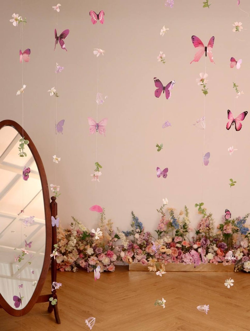 Pu&ograve; includere: Un muro rosa con una ghirlanda floreale con farfalle appese. La ghirlanda &egrave; fatta di farfalle e fiori di carta. La ghirlanda &egrave; appesa davanti a uno specchio.