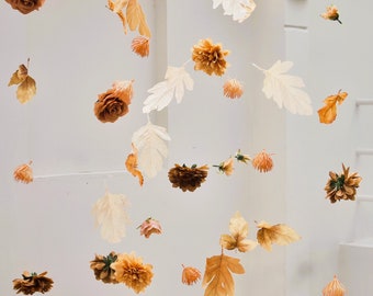 May include: An overhead shot of hanging floral decorations. The arrangement features various paper flowers and leaves in shades of brown, orange, and cream. The backdrop is a white wall with architectural details.