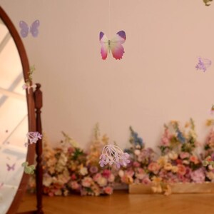 Pu&ograve; includere: Una ghirlanda decorativa con fiori viola e bianchi e farfalle appese a un muro bianco. La ghirlanda &egrave; fatta di carta ed &egrave; infilata su un filo trasparente. La ghirlanda &egrave; appesa davanti a un muro di fiori.