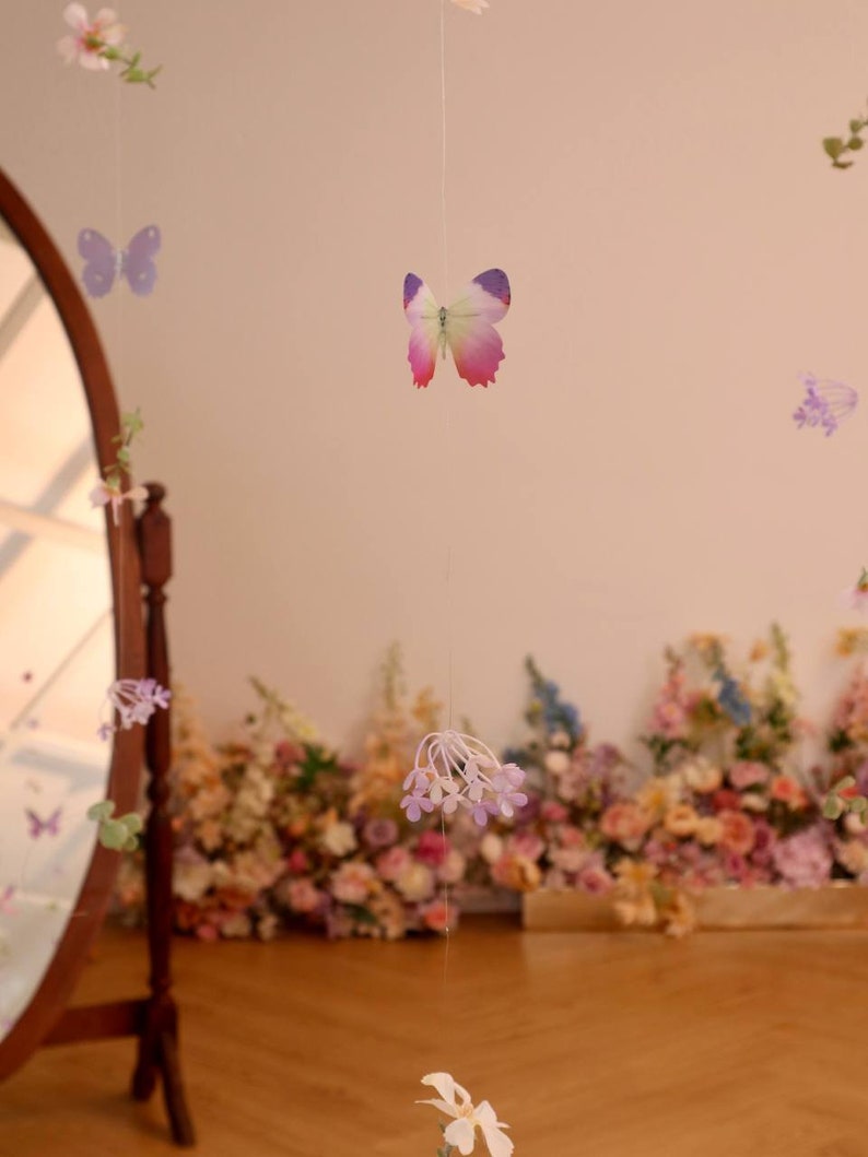 Pu&ograve; includere: Una ghirlanda decorativa con fiori viola e bianchi e farfalle appese a un muro bianco. La ghirlanda &egrave; fatta di carta ed &egrave; infilata su un filo trasparente. La ghirlanda &egrave; appesa davanti a un muro di fiori.