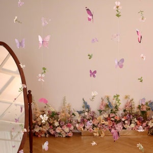 Pu&ograve; includere: Un set di ghirlande di farfalle e fiori di colore viola e rosa appese a un muro bianco. Le ghirlande sono fatte di carta e sono infilate su un filo trasparente. Le farfalle sono di diverse dimensioni e forme, e i fiori sono bianchi e rosa. Le ghirlande sono appese davanti a un muro di fiori rosa e bianchi.