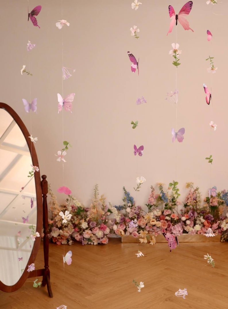 Pu&ograve; includere: Un set di ghirlande di farfalle e fiori di colore viola e rosa appese a un muro bianco. Le ghirlande sono fatte di carta e sono infilate su un filo trasparente. Le farfalle sono di diverse dimensioni e forme, e i fiori sono bianchi e rosa. Le ghirlande sono appese davanti a un muro di fiori rosa e bianchi.