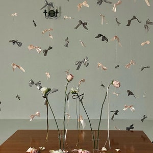 Może przedstawiać: Nakrycie stołu z czterema przezroczystymi szklanymi wazonami wypełnionymi r&oacute;żowymi i białymi kwiatami. Wazy są ozdobione szarymi i r&oacute;żowymi wstążkami. St&oacute;ł jest drewniany i ma jasnobrązowe wykończenie.