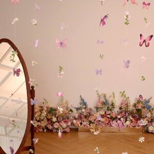 Pu&ograve; includere: Un set di ghirlande di farfalle e fiori rosa e viola appese a un muro con uno specchio e un'arrangiamento floreale in basso.