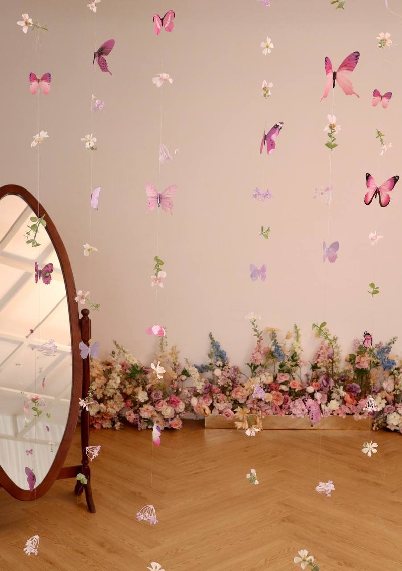 Pu&ograve; includere: Un set di ghirlande di farfalle e fiori rosa e viola appese a un muro con uno specchio e un'arrangiamento floreale in basso.