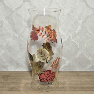 May include: A clear glass hurricane vase featuring a hand-painted design of autumn leaves in shades of red, orange, brown, and yellow. The exterior surface is decorated with scattered gold glitter accents throughout the leaf pattern.