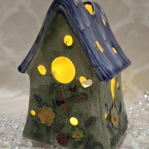 Peut inclure: Un bougeoir en céramique en forme de maison avec un toit bleu et des murs verts. La maison a des fenêtres découpées et un motif de libellule. Le bougeoir est allumé et brille.