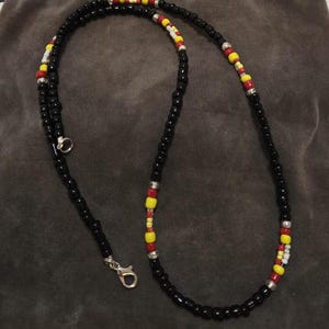 Puede incluir: Collar de cuentas negras con cuentas de acento rojas, amarillas y blancas. El collar tiene un cierre plateado y un pequeño anillo plateado. Las cuentas son pequeñas y redondas, y el collar se muestra sobre una superficie gris oscuro.