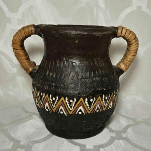 Op de afbeelding: Een donkerbruine keramische vaas met twee geweven handvatten. De vaas heeft een decoratieve band van witte, gele en rode geometrische patronen. Het oppervlak heeft gesneden verticale lijnen en een textuur.