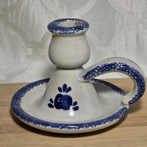Op de afbeelding: Een keramische kandelaar met een witte basis en een blauw bloemmotief. De houder heeft een handvat en een cirkelvormige basis met een blauwe stippelrand. De bovenrand en het handvat zijn ook versierd met blauwe stippen.