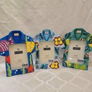 Puede incluir: Tres marcos de fotos decorativos con forma de camisas hawaianas. Cada marco mide 8,9 x 12,7 cm y presenta un diseño único con peces tropicales, flores y hojas. Los marcos son en tonos de azul, verde y amarillo.