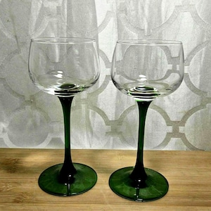 May include: Two clear wine glasses with green stems and bases. The glasses have a rounded bowl shape and sit on a wooden surface. The stems and bases are a deep, forest green color.