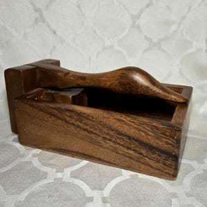 May include: A wooden box with a curved handle. The box is rectangular with a dark brown finish, and the handle is a lighter shade of brown. The handle is curved and appears to be attached to the top of the box.