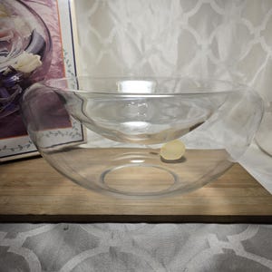 May include: A clear glass bowl with a unique, layered design. The bowl has a larger outer layer and a smaller inner layer, creating a visually interesting shape. The bowl sits on a wooden surface.