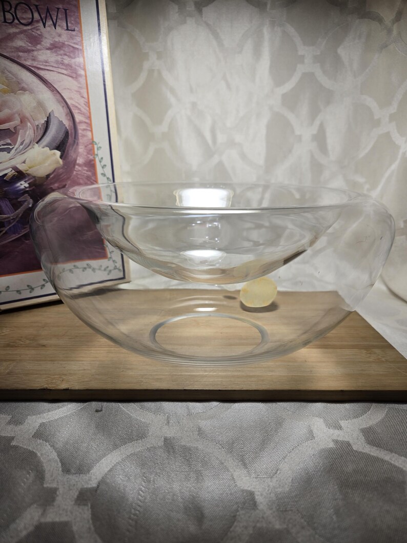 May include: A clear glass bowl with a unique, layered design. The bowl has a larger outer layer and a smaller inner layer, creating a visually interesting shape. The bowl sits on a wooden surface.