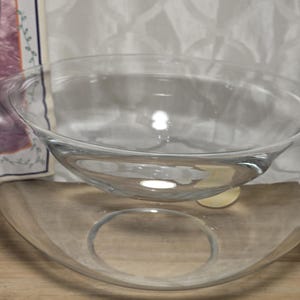 May include: Two clear glass bowls, one nested inside the other, sit on a wooden surface. The bowls are transparent, allowing light to pass through. The background is a white patterned fabric.