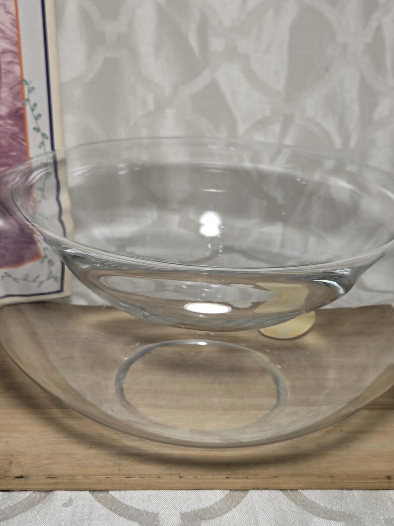 May include: Two clear glass bowls, one nested inside the other, sit on a wooden surface. The bowls are transparent, allowing light to pass through. The background is a white patterned fabric.