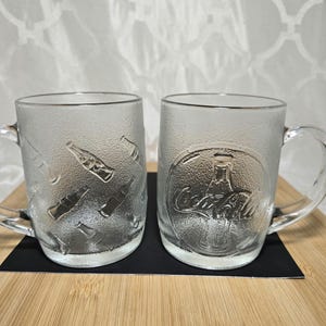 Könnte beinhalten: Zwei transparente Glasbecher mit Henkeln. Ein Becher zeigt geprägte Coca-Cola-Flaschenmotive, während der andere das Coca-Cola-Logo hat. Die Becher stehen auf einer Holzoberfläche mit schwarzem Untergrund.