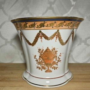 May include: A white ceramic vase with a flared shape, featuring gold trim and decorative orange and gold floral and garland designs. The vase has a base diameter of approximately 4 inches.