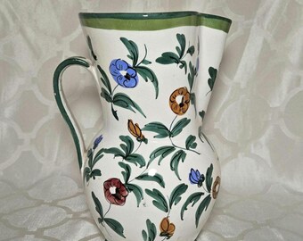 Op de afbeelding: Witte keramische kan met een groene handgreep en voet, versierd met kleurrijke bloemen- en bladerpatronen. De kan heeft een geschulpte rand en een groene rand, waardoor het een decoratief en functioneel stuk is.