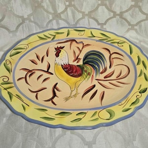 Puede incluir: Plato ovalado de cerámica con un diseño de gallo pintado a mano. El plato presenta un borde amarillo con detalles de hojas verdes y un borde azul. El gallo es amarillo, rojo y azul, con detalles de hojas marrones.