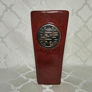 May include: A rectangular vase in a rich, deep red glaze. A circular, bronze-toned emblem is centered on the front. The vase has a slightly tapered shape and a glossy finish, suitable for home decor.