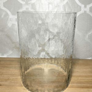 May include: A clear glass cylinder vase featuring a textured, hammered surface finish. The glass has a uniform cylindrical shape with an open top and a flat base, creating a translucent appearance that allows light to pass through the patterned walls.