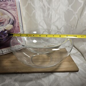 May include: A clear glass bowl with a unique design, approximately 10 inches in diameter, sits on a wooden board. The bowl has a smaller, elevated inner section and a circular opening. A measuring tape is placed across the bowl.