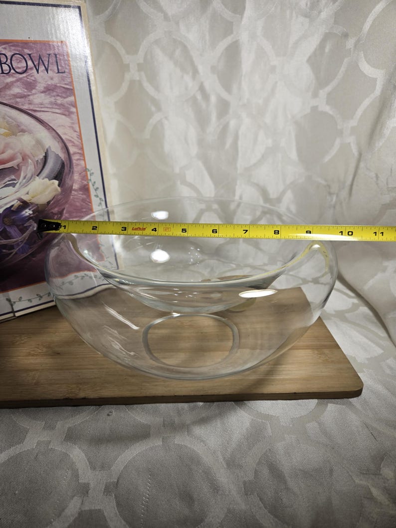 May include: A clear glass bowl with a unique design, approximately 10 inches in diameter, sits on a wooden board. The bowl has a smaller, elevated inner section and a circular opening. A measuring tape is placed across the bowl.