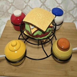 Peut inclure: Un porte-condiments en céramique en forme de sandwich avec quatre petits pots à condiments. Le sandwich est jaune, vert et rouge. Les pots sont jaunes, orange, rouge et blanc. Le support est sur une surface en bois.