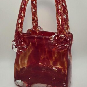 Peut inclure: Un sac à main en verre rouge avec une poignée torsadée. Le sac a un aspect marbré et moucheté.