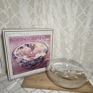 May include: A clear glass floating candle bowl with a box that reads "Floating Candle Bowl". The box shows the bowl filled with floating candles and flowers. The bowl is on a wooden surface.