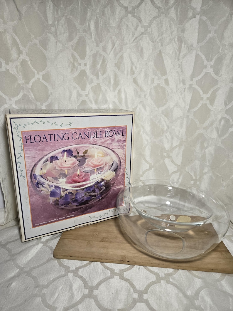 May include: A clear glass floating candle bowl with a box that reads "Floating Candle Bowl". The box shows the bowl filled with floating candles and flowers. The bowl is on a wooden surface.