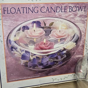 May include: A clear glass floating candle bowl with three lit rose-shaped candles. The bowl contains pink, white, and purple artificial flowers. The box reads "FLOATING CANDLE BOWL".