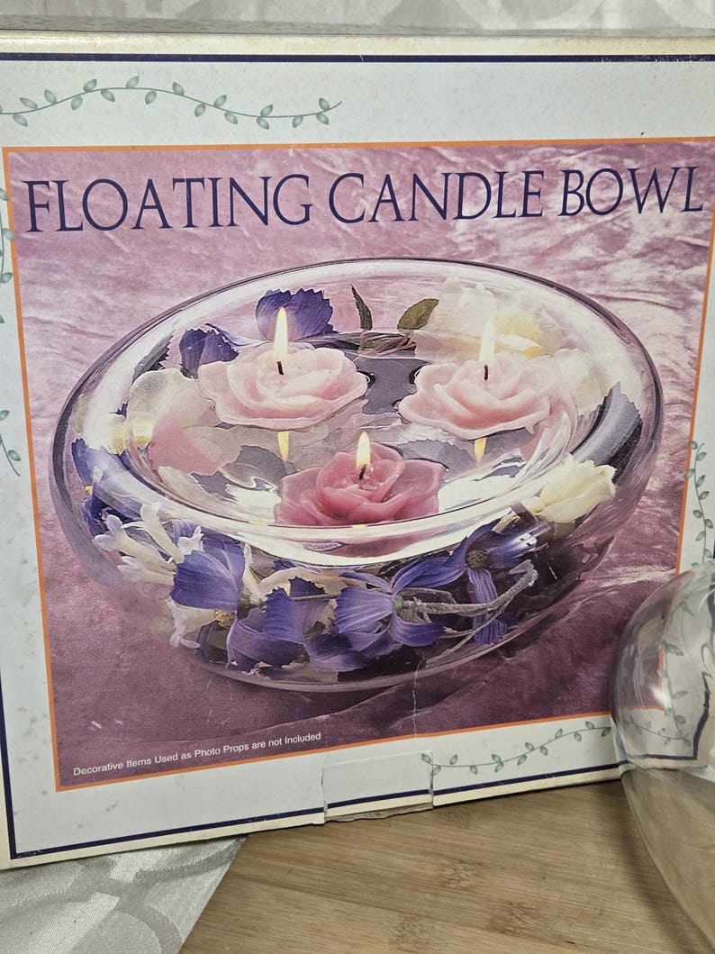 May include: A clear glass floating candle bowl with three lit rose-shaped candles. The bowl contains pink, white, and purple artificial flowers. The box reads "FLOATING CANDLE BOWL".