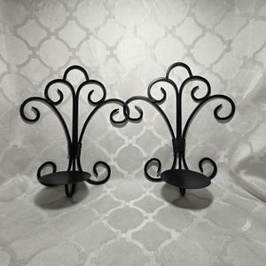May include: A pair of black metal wall sconces featuring a symmetrical scrollwork design with a central vertical spine and a circular platform base for holding candles. The metal structure is composed of curved wire elements in a fleur-de-lis shape.
