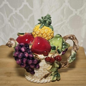 Può includere: Una teiera in ceramica a forma di cestino traboccante di frutta colorata. La teiera presenta un ananas, mele rosse, uva, ciliegie e foglie verdi. Il manico e il beccuccio sono progettati per assomigliare a rami.