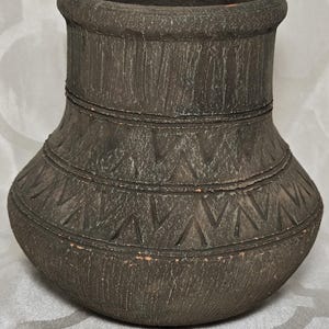 Op de afbeelding: Een donkerbruine, getextureerde keramische vaas met een brede opening en een afgeronde basis. De vaas heeft geëtste geometrische patronen en horizontale banden. Het algehele ontwerp heeft een rustieke, handgemaakte uitstraling.