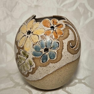 May include: A round ceramic vase with a speckled beige base and a cream-colored top. The vase is decorated with colorful flowers in yellow, orange, and blue, with brown accents. The flowers have black centers.